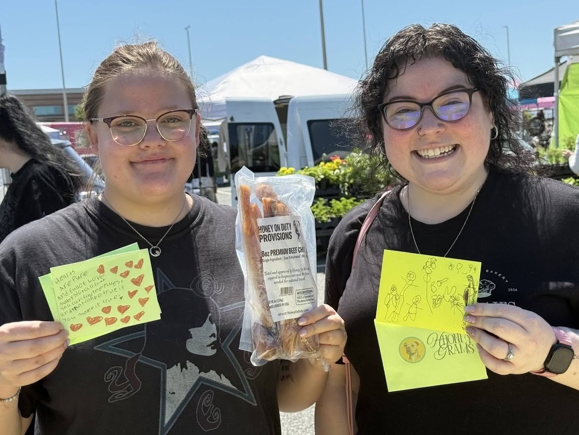Two women holding Honey's Dog Treats and yellow cards at a community event
